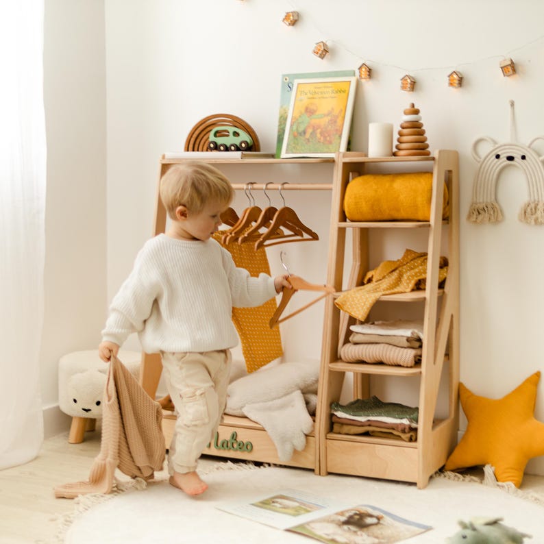 May include: A light-colored wooden clothing rack with a hanging rod, multiple shelves, and the name Mateo in green lettering. The rack holds wooden hangers, folded garments, a yellow blanket, and a stack of books. A small wooden car sits on top.