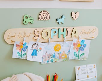 以下が含まれることがあります： パステルカラーの文字で「SOPHIA」と書かれた木製のディスプレイボード。子供たちの絵が飾られています。トラクター、虹、ユニコーン、ハートなどの装飾的な木製シェイプがあります。「Look What Made」というテキストも表示されています。