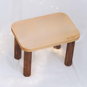 Puede incluir: Un taburete peque&ntilde;o de madera con una superficie rectangular de madera clara natural con esquinas redondeadas y cuatro patas cil&iacute;ndricas de madera te&ntilde;idas de marr&oacute;n oscuro. La parte superior tiene un grabado de marca tenue a la derecha.