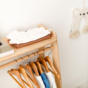 May include: A light-colored wooden clothing rack featuring a top shelf with a folded white shirt with small orange dots and a solid brown shirt. Below, five wooden hangers hold folded garments in white, blue, and textured white fabric.