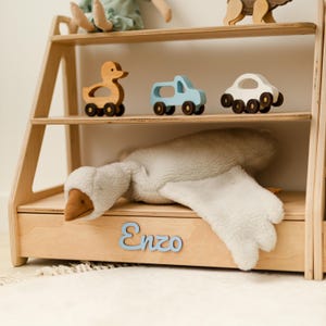May include: A light-colored wooden bookshelf with three tiers. The middle shelf holds a wooden duck, a light blue truck, and a white car. The bottom shelf features a plush white goose toy and the name Enzo in light blue script lettering.