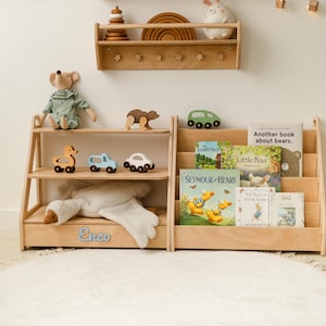 May include: A light-colored wooden bookshelf and a matching book display rack with three tiers. The bookshelf features the name Enzo in blue script. The display rack holds several children's books, including titles like Seymour & Henry and Little Baa.