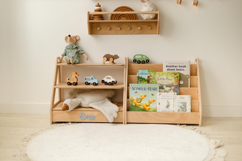 May include: A light-colored wooden bookshelf and a matching book display rack with three tiers. The bookshelf features the name Enzo in blue script. The display rack holds several children's books, including titles like Seymour & Henry and Little Baa.