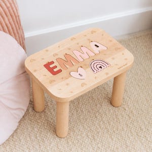 Puede incluir: Un taburete de madera clara con una superficie tipo rompecabezas que muestra el nombre EMMA en tonos degradados de rosa y terracota. La superficie incluye motivos grabados de arco&iacute;ris y soles, una pieza con forma de coraz&oacute;n y otra de arco&iacute;ris.
