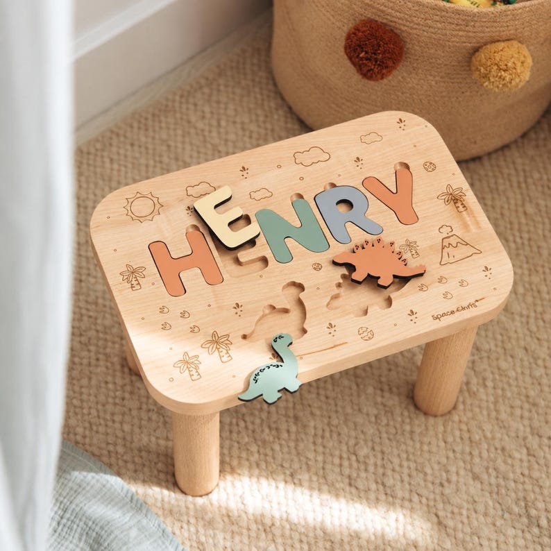 Puede incluir: Un taburete de madera clara con una superficie de rompecabezas que muestra el nombre HENRY en letras de colores pastel. La superficie incluye motivos grabados de dinosaurios, palmeras, sol y volc&aacute;n, con una pieza de rompecabezas verde.