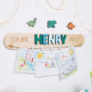 Op de afbeelding: Een houten displaybord met de naam "HENRY" in turquoise en blauwe letters, en de woorden "LOOK WHAT MADE" in lichtbruine letters. Het bord heeft een touwtje met wasknijpers met kindertekeningen en dinosaurussen en beren.