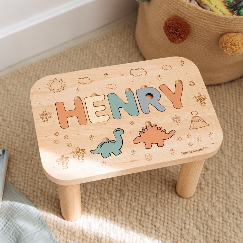 Puede incluir: Un taburete de madera de color claro con el nombre HENRY en letras recortadas de tonos pastel. La superficie est&aacute; decorada con ilustraciones grabadas de un sol, nubes, palmeras, un volc&aacute;n y dos dinosaurios en azul y naranja.