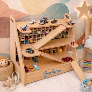 May include: A light-colored wooden toy car storage shelf with multiple compartments, ramps, and levels. The name Graham is written in blue script on the front. The unit holds various miniature toy vehicles, including cars, trucks, and planes.