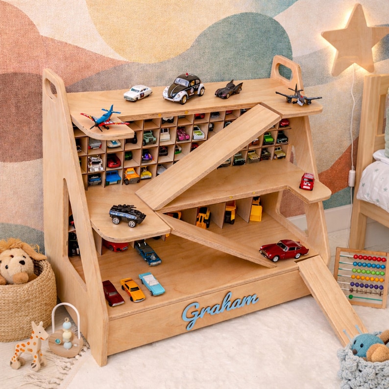 May include: A light-colored wooden toy car storage shelf with multiple compartments, ramps, and levels. The name Graham is written in blue script on the front. The unit holds various miniature toy vehicles, including cars, trucks, and planes.
