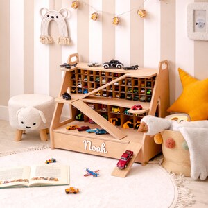 May include: A light-colored wooden toy car storage shelf with a grid of compartments, a slide-out ramp, and a drawer featuring the name Noah in white script lettering. The unit holds numerous small toy vehicles and airplanes on its various levels.