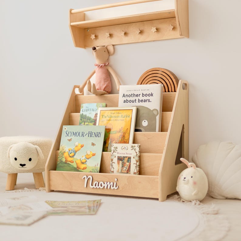 May include: A light-colored wooden bookshelf with four tiered shelves, featuring the name Naomi in white script on the front. The shelf holds several children's books, including titles like Seymour and Henry and Another book about bears.