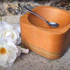 May include: A square wooden bowl with a smooth, light-toned upper section and a textured, darker-toned base. A small, polished silver-coloured metal spoon rests inside the bowl. Two white flowers with yellow centres are positioned next to the bowl.