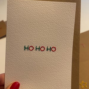 Può includere: Un biglietto di auguri bianco strutturato con la scritta ricamata "HO HO HO" in filo verde e rosso. Il biglietto è all'interno di una busta marrone. L'immagine mostra un primo piano del biglietto e della busta.