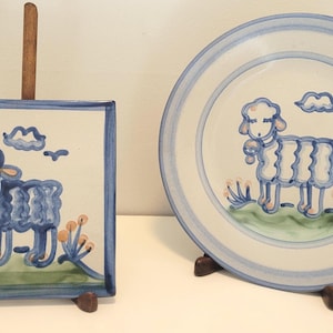 May include: A square ceramic plate and a round ceramic plate, both featuring a hand-painted blue sheep design on a white background. Each plate displays a sheep standing on a green patch with small orange flowers, a blue cloud, and a bird.
