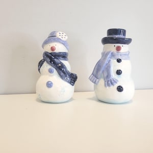 May include: Two ceramic snowman figurines with glossy white bodies and blue accents. The left snowman wears a blue hat and dark blue scarf. The right snowman wears a dark blue top hat and a light blue scarf with three dark blue buttons on its front.