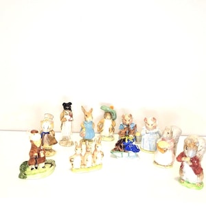May include: A collection of twelve small ceramic animal figurines, including rabbits and squirrels dressed in colorful clothing. The figures are painted in various pastel and earth tones, depicting characters in different poses and outfits.