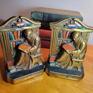 Peut inclure: Paire de serre-livres de couleur bronze représentant un moine lisant devant une bibliothèque. Les serre-livres ont un cadre doré et une base noire. La bibliothèque est remplie de livres colorés. Le moine est assis sur un petit tabouret et tient un livre orange.
