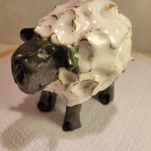 May include: A ceramic sheep figurine featuring a dark, metallic-toned head and legs contrasted with a thick, glossy white glazed body. The surface of the wool has a textured, uneven appearance with visible brown undertones beneath the white glaze.