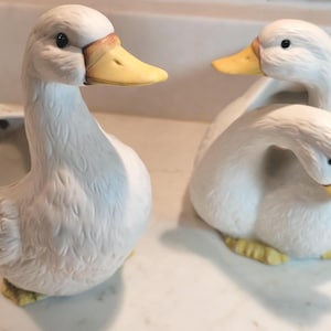 May include: Two white ceramic duck figurines with yellow beaks and feet. One duck is standing upright with an open hollow back, while the other duck is sitting with its head tucked down. Both feature textured feather details and black eyes.