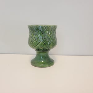 May include: A green ceramic goblet featuring a textured diamond pattern across the bowl. The vessel has a smooth, flared base and a glossy finish with variations in color intensity, creating a mottled appearance across the surface of the piece.