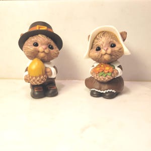 Könnte beinhalten: Zwei kleine Tierfiguren in historischer Kleidung. Eine Figur trägt einen dunkelbraunen Hut und hält eine gelbe Eichel. Die andere Figur trägt eine weiße Haube und hält einen Korb mit orangefarbenen Blumen. Beide haben braunes Fell.