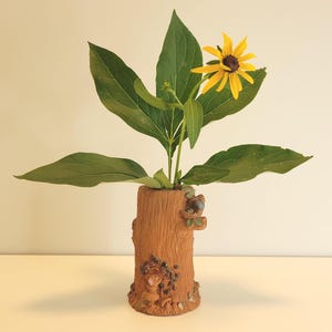 May include: A brown ceramic vase shaped like a tree stump with textured bark details and small sculpted figures of a mouse and mushrooms at the base. A single yellow flower with a dark center and several green leaves are arranged inside the vase.