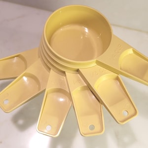 Può includere: Un set di cinque misurini in plastica giallo pallido impilati a ventaglio. Ogni manico presenta un piccolo foro circolare all'estremità ed è impresso con indicazioni di volume tra cui 1/4 cup, 1/3 cup, 1/2 cup, 2/3 cup e 1 cup.