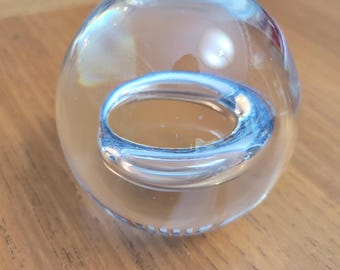 May include: A clear glass paperweight shaped as a sphere with a smooth, polished surface. Inside the transparent glass, there is a distinct, hollow, ring-like circular cavity that creates a reflective, distorted visual effect within the orb.