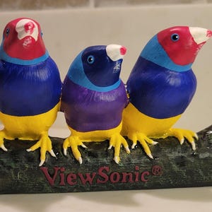 Puede incluir: Una figura decorativa con tres pájaros coloridos posados sobre un tronco texturizado de color verde oscuro. Los dos pájaros exteriores tienen cabezas rojas, cuellos azules y cuerpos amarillos, mientras el central tiene cabeza azul.