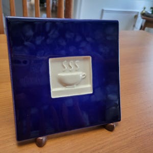 May include: A square, deep blue ceramic tile featuring a raised, cream-coloured coffee cup motif. The design includes steam lines above the cup. The tile is displayed on a wooden easel.