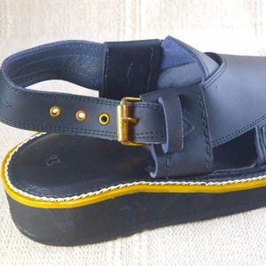 May include: Close-up of a black leather sandal with a thick sole. The sandal features a strap with a gold-colored buckle and decorative gold rivets. The sole has a yellow and white trim.