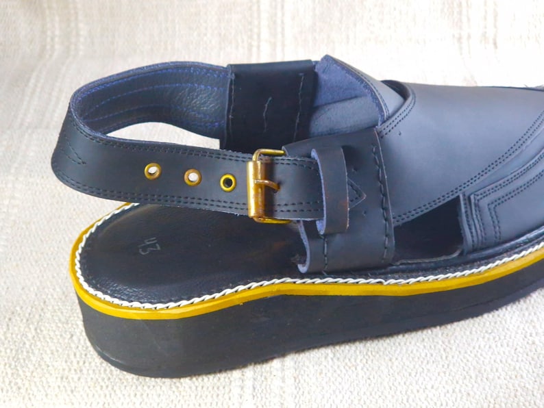 May include: Close-up of a black leather sandal with a thick sole. The sandal features a strap with a gold-colored buckle and decorative gold rivets. The sole has a yellow and white trim.