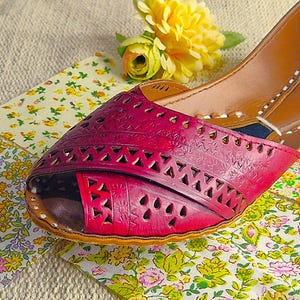 May include: Close-up of a burgundy leather shoe with intricate cut-out designs. The shoe is set against a backdrop of floral patterned fabric and a yellow flower, showcasing a blend of textures and colors.