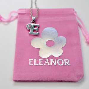 May include: A pink velvet pouch with a silver charm necklace featuring the letter "E" with a small flower detail. The pouch has a silver flower and the word "ELEANOR" printed on it.