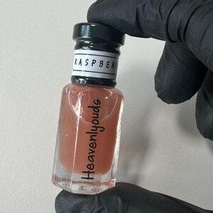 Pode incluir: Uma pequena garrafa de vidro com uma tampa preta e um líquido castanho no seu interior. A garrafa tem um rótulo que diz "Raspberry" e "Heavenlyjouds".