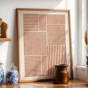 Op de afbeelding: Een keukeninterieur met een ingelijste abstracte print in bruine en crèmetinten. Houten planken tonen borden, kommen en keukengerei. Decoratieve potten met blauwe bloempatronen en een bruine vaas staan op het aanrecht.