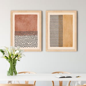 May include: Two framed wall art prints featuring abstract geometric designs. The left print displays a terracotta block above dark zigzag lines. The right print shows horizontal dark stripes next to a solid mustard yellow block. Light wood frames.