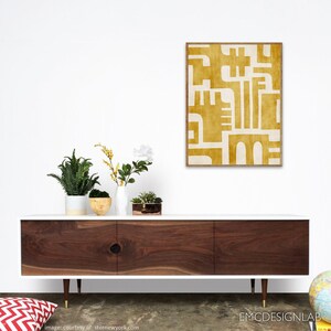 May include: A white mid-century modern dresser with three drawers and gold legs. The dresser has a wood grain top and is decorated with a yellow and white abstract art print.