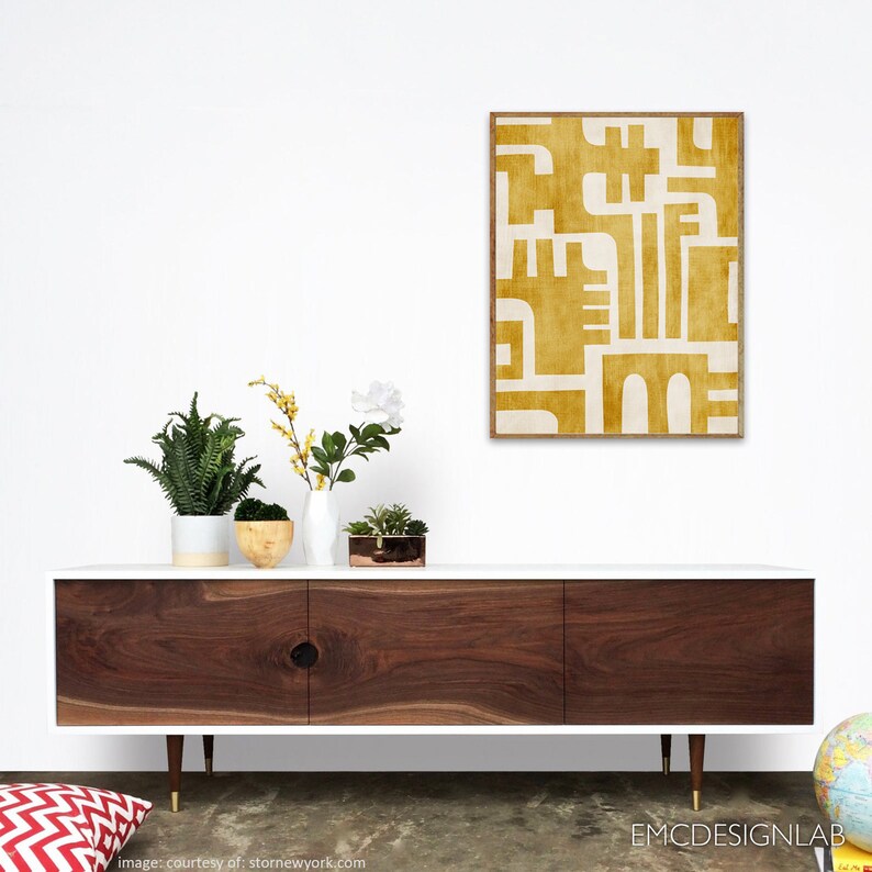 May include: A white mid-century modern dresser with three drawers and gold legs. The dresser has a wood grain top and is decorated with a yellow and white abstract art print.