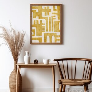 May include: A framed print with a geometric design in a mustard yellow color on a white background. The print is hanging on a white wall above a wooden table with a vase, a mug, and a small wooden object.