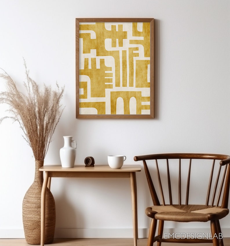 May include: A framed print with a geometric design in a mustard yellow color on a white background. The print is hanging on a white wall above a wooden table with a vase, a mug, and a small wooden object.