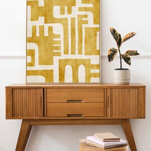 May include: A mid-century modern console table with two drawers and slatted cabinet doors. The table is made of wood and has a warm brown finish. A yellow abstract painting hangs above the table. A small wooden stool sits in front of the table with a stack of books on top.