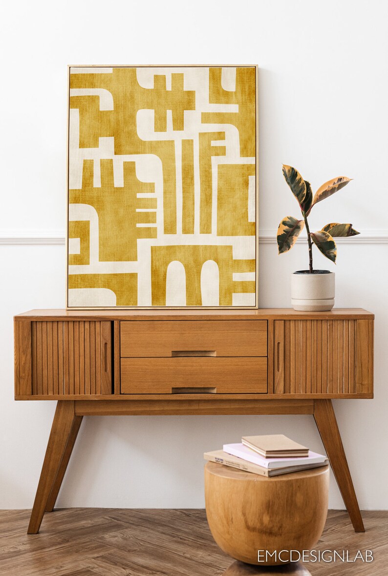 May include: A mid-century modern console table with two drawers and slatted cabinet doors. The table is made of wood and has a warm brown finish. A yellow abstract painting hangs above the table. A small wooden stool sits in front of the table with a stack of books on top.