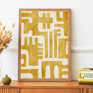 May include: A framed abstract print with a geometric design in shades of yellow and white. The print is set against a white wall and a wooden table with a vase of dried flowers and a green ceramic vase.