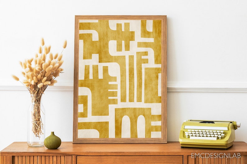 May include: A framed abstract print with a geometric design in shades of yellow and white. The print is set against a white wall and a wooden table with a vase of dried flowers and a green ceramic vase.