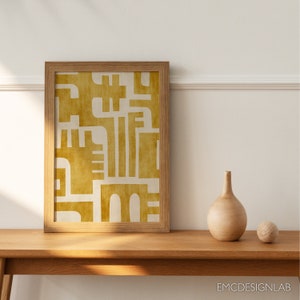 May include: A framed print with a geometric design in shades of yellow and white. The print is hanging on a white wall above a wooden table with a vase and a ball on it.