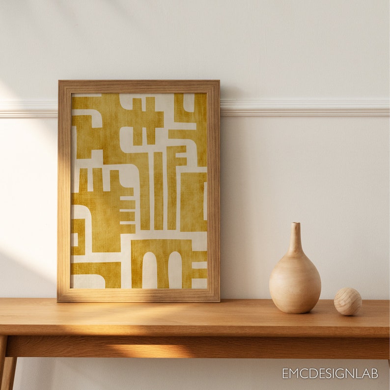 May include: A framed print with a geometric design in shades of yellow and white. The print is hanging on a white wall above a wooden table with a vase and a ball on it.