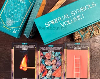 May include: A deck of Spiritual Symbols Volume I oracle cards in a teal box, with three cards displayed face up. The cards feature images of a lit match, a pile of colorful stones, and a ladder, each with descriptive text and meanings printed.
