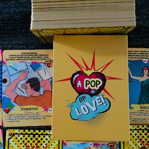 Peut inclure: Un jeu de cartes avec une bo&icirc;te jaune orn&eacute;e d'un c&oelig;ur rouge et du texte A POP OF LOVE. Les cartes pr&eacute;sentent des illustrations de style bande dessin&eacute;e, incluant une personne au repos, un personnage au chapeau et divers motifs de c&oelig;urs.
