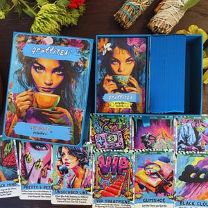 May include: A blue box set of GraffiTEA tarot cards. The box features a woman with floral accents holding a teacup. The cards have colorful illustrations and text, including "Vibe About It" and "VIP Treatment."
